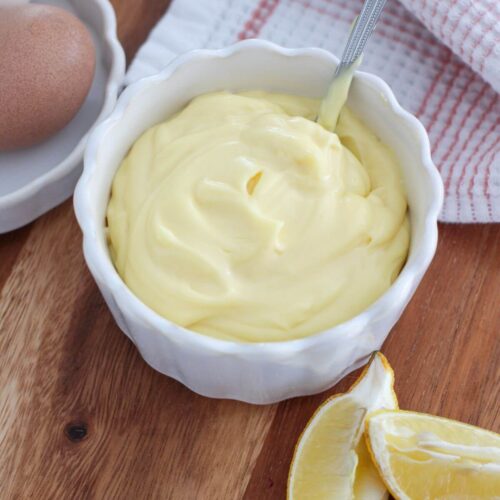 overhead view of mayo in a white bowl on a cutting board with lemon slices and eggs next to it in a white dish with a pink and white towel