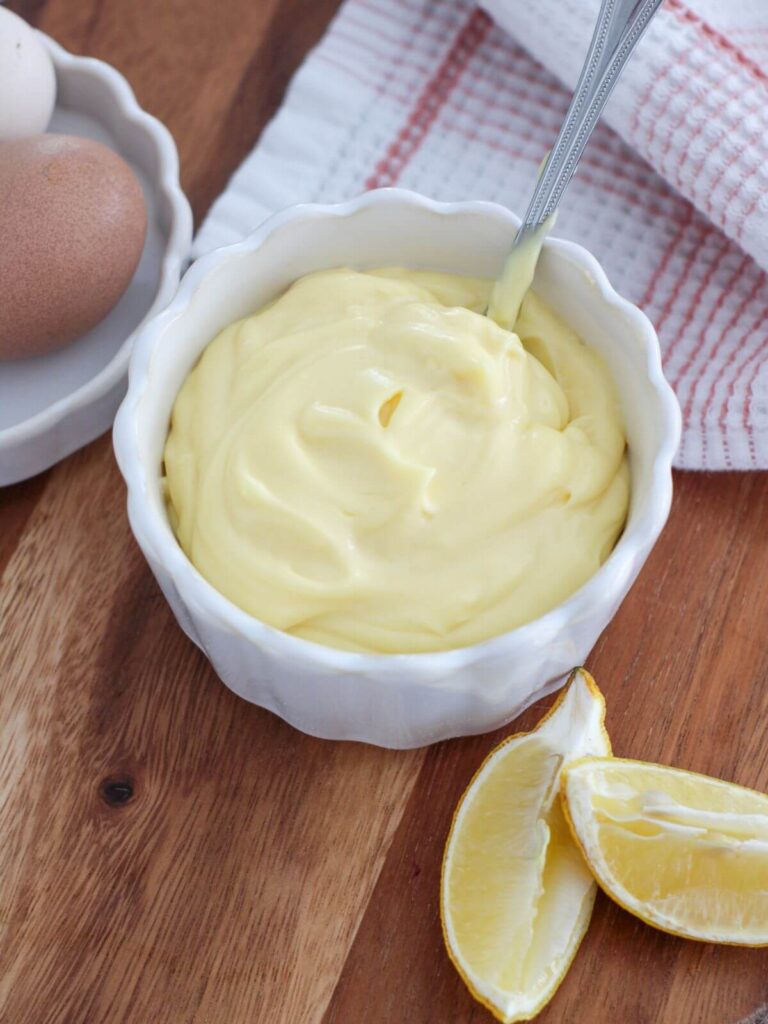 overhead view of mayo in a white bowl on a cutting board with lemon slices and eggs next to it in a white dish with a pink and white towel 