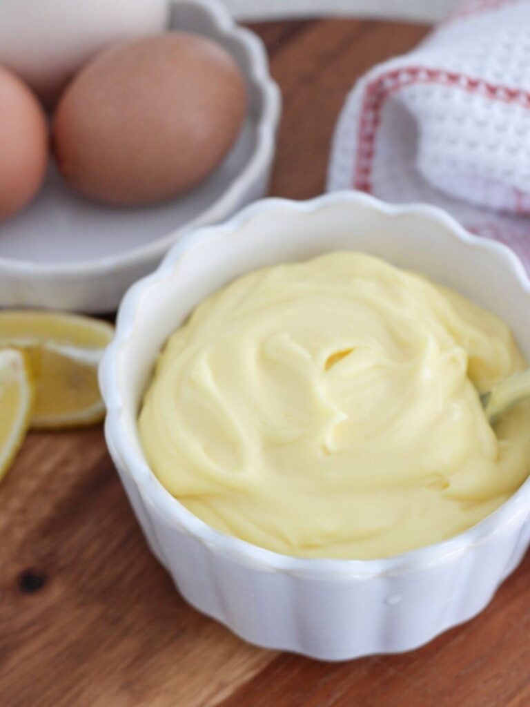 Side view of mayo in a white dish on a cutting board with lemon wedges and eggs on a white plate with a towel next to it