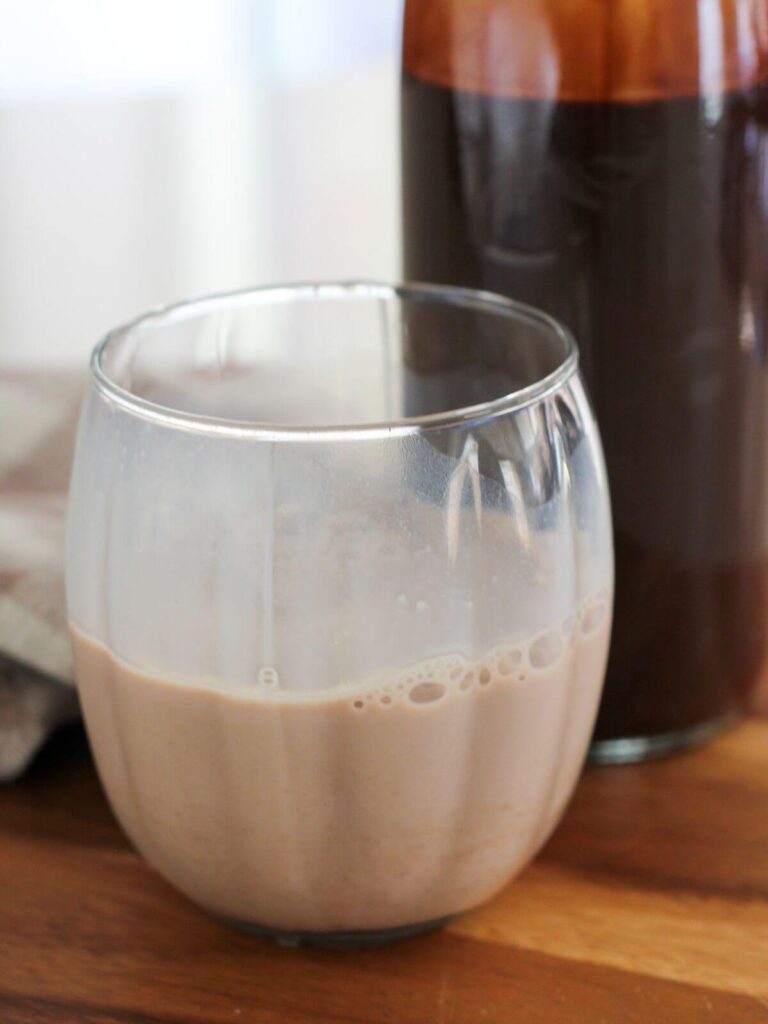 Milk with chocolate syrup in it in a clear glass with chocolate syrup bottle behind it on a cutting board with a plaid napkin