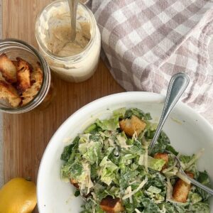 a Caesar salad made in a white bowl with a spoon. Lemons next to it and croutons and Caesar dressing in a mason jar with a plaid towel next to it all on a cutting board.
