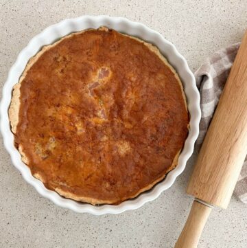 ham and cheese quiche in a white pie pan with a rolling pin and plaid towel next to it