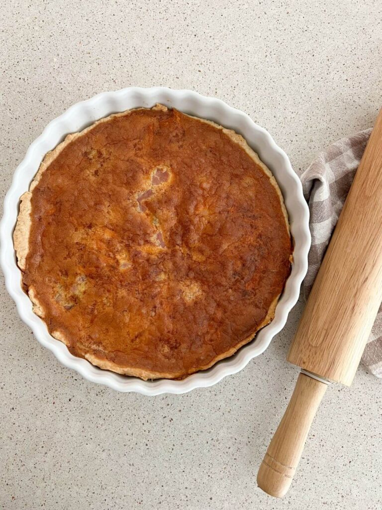 ham and cheese quiche in a white pie pan with a rolling pin and plaid towel next to it