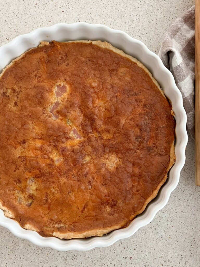 ham and cheese quiche in a white pie pan with a rolling pin and plaid towel next to it