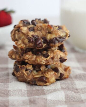 3 Simple Banana Breakfast Cookie stacked on top of each other on a plaid napkin with milk in a clear glass and strawberries in a white bowl in the background.