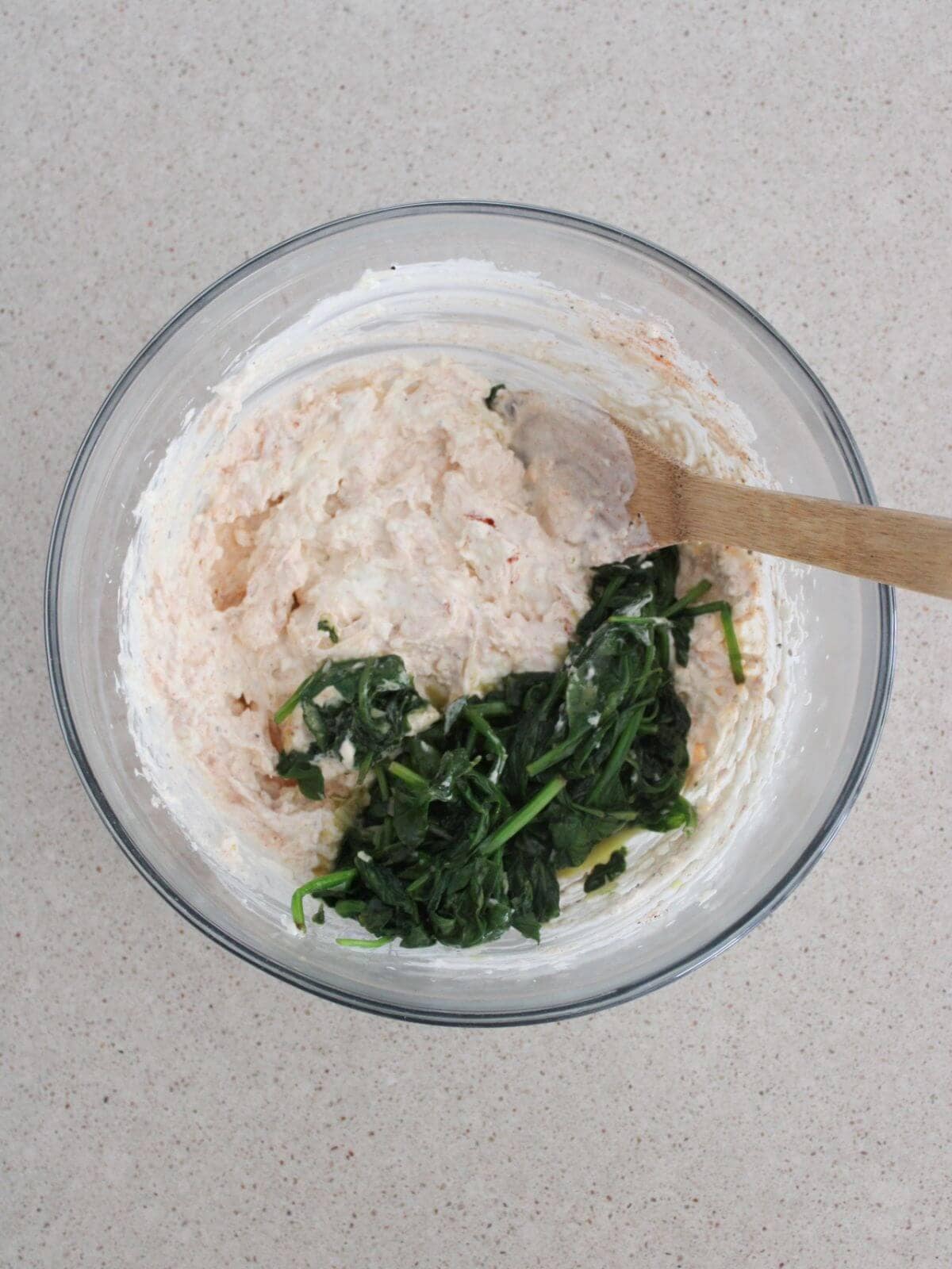 spinach being folded into the mixture in a glass bowl with a wooden spoon.