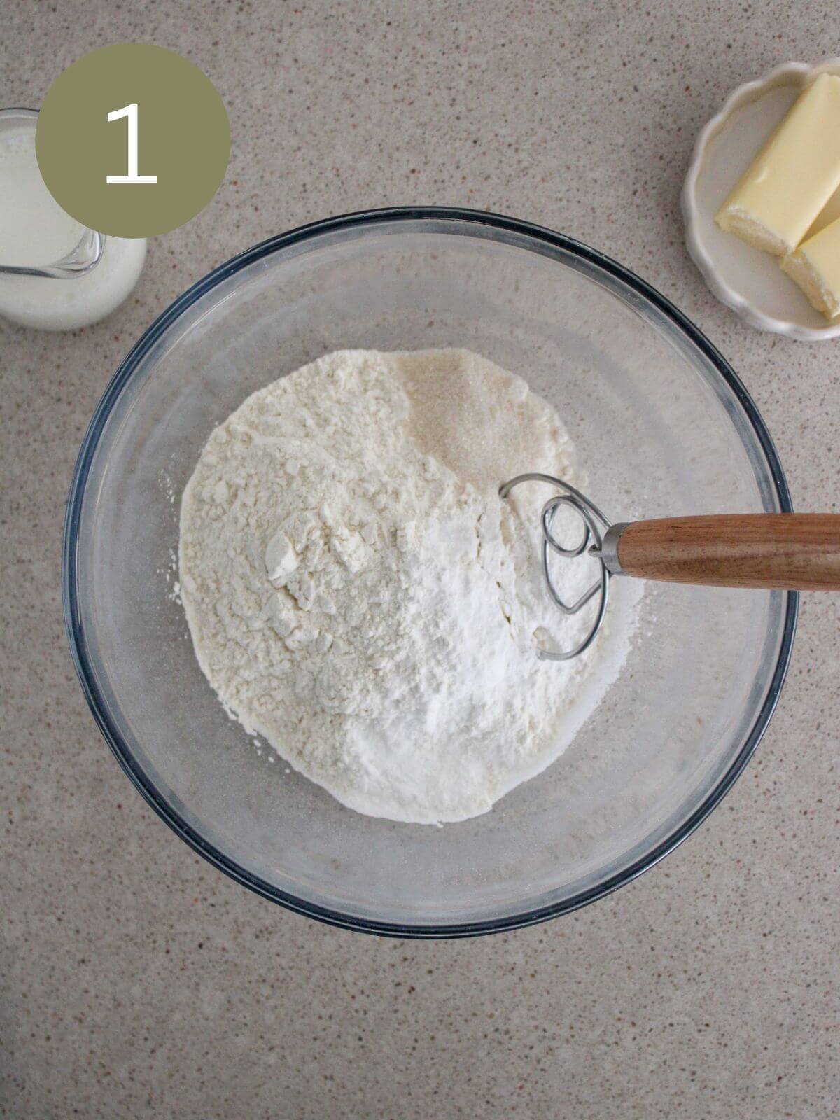 Flour, salt, baking powder and sugar mixed in a clear glass bowl with a dough whisk with milk and butter off to the side