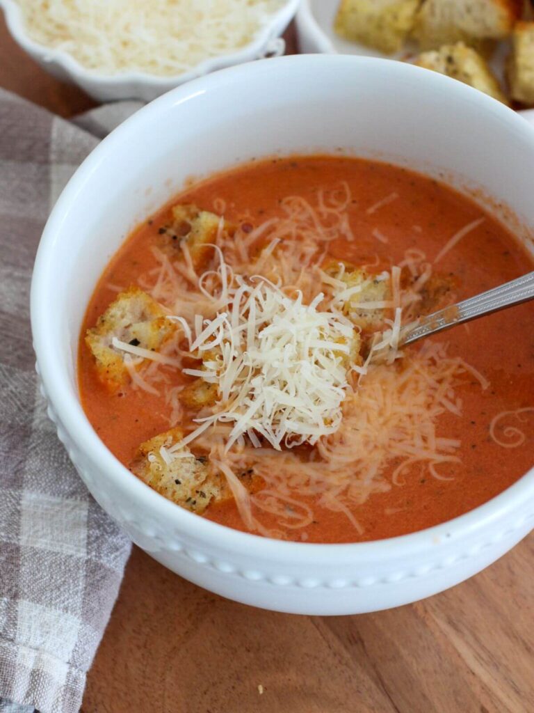 Side view of the tomato soup in a white bowl on a cutting board with a plaid napkin with croutons and parmesan in small white bowls above