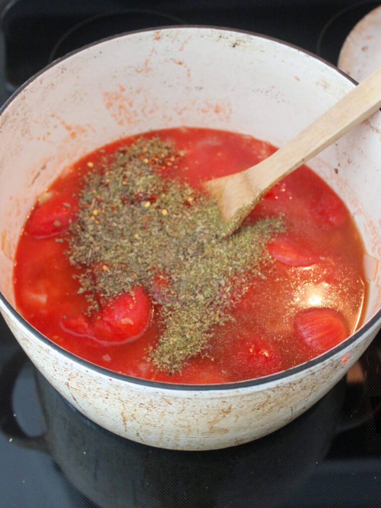 whole, peeled tomatoes with basil, oregano and red pepper flakes in a white dutch oven with a wooden spoon