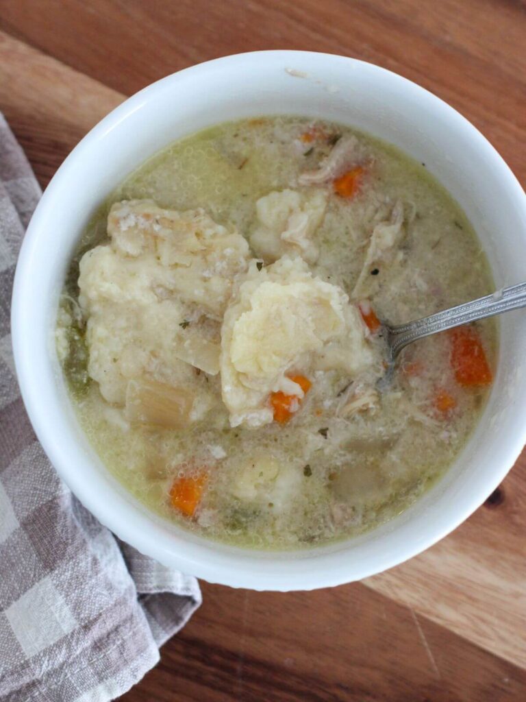 chicken and dumplings in a white bowl and a silver spoon in it on a cutting board with a plaid napkin