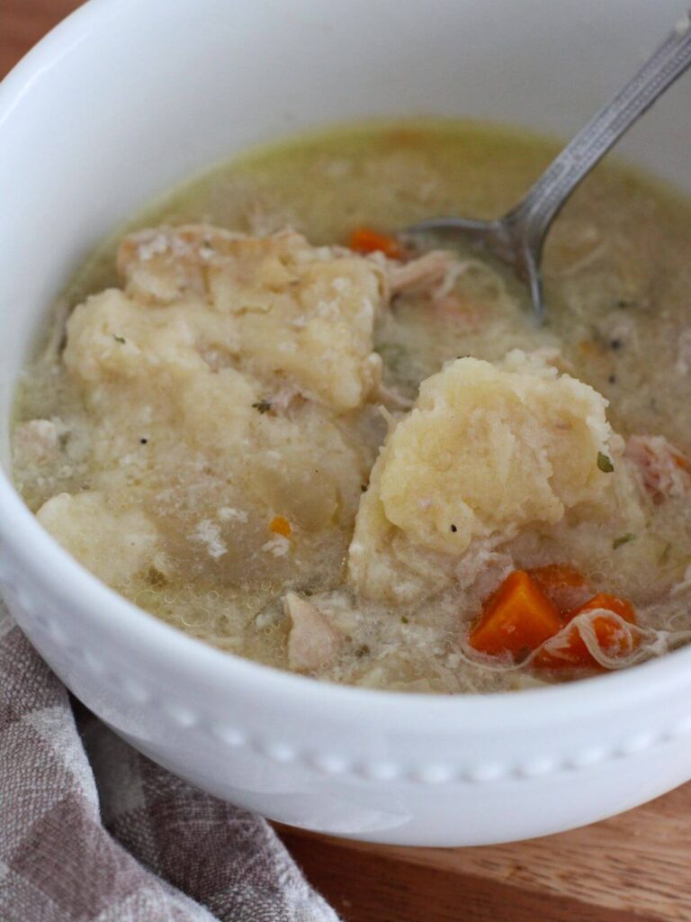 Slow cooker chicken and dumplings up close in a white bowl with a silver spoon a cutting board with a plaid napkin