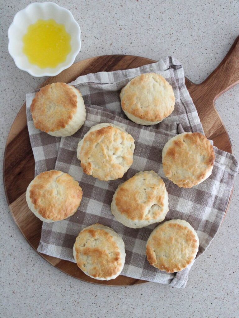 Homemade biscuits scattered on a plaid napkin on top of a cutting board with a little white bowl of butter next to it