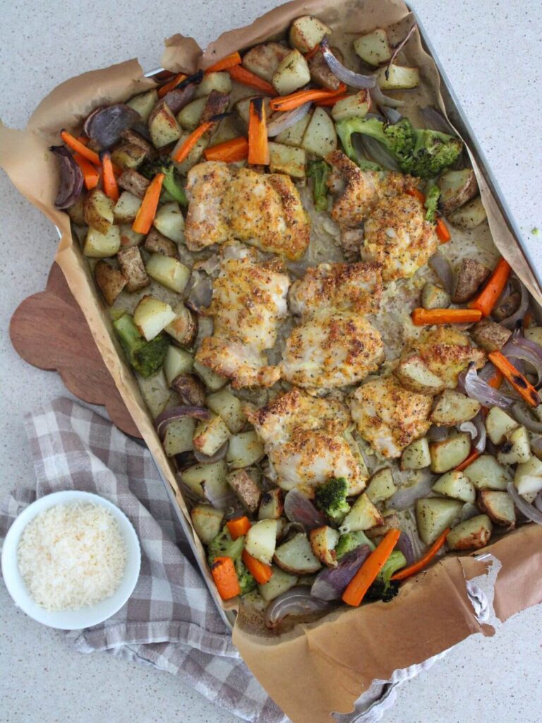 Easy Sheet Pan Parmesan Chicken with veggies and potatoes on a sheet pan lined with parchment paper on top a cutting board with a plaid napkin with a white bowl with parmesan in it.