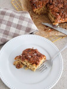 Simple Maple Cinnamon Snacking Cake on a plate cut with the rest of the cake behind it on parchment paper and a golden cooling rack and a silver knife an a plaid napkin next to it.