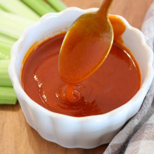 hot sauce in a white ramekin with a silver spoon on a cutting board with celery and plaid napkin