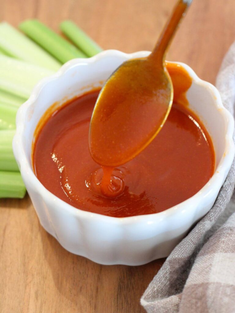 hot sauce in a white ramekin with a silver spoon on a cutting board with celery and plaid napkin