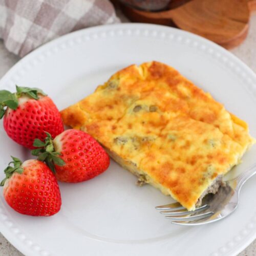a piece of the egg and sausage bake on a white plate with 3 strawberries next to it and a plaid napkin with the rest of the bake in the background