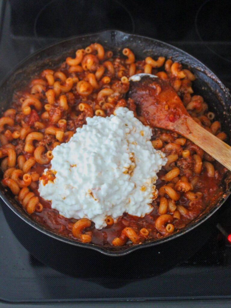 adding the cottage cheese into the pasta mixture in a cast iron skillet with a wooden spoon