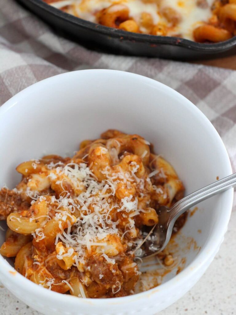 pasta bake spooned into a white bowl with parmesan on top with the cast iron skillet in the back with a plaid napkin