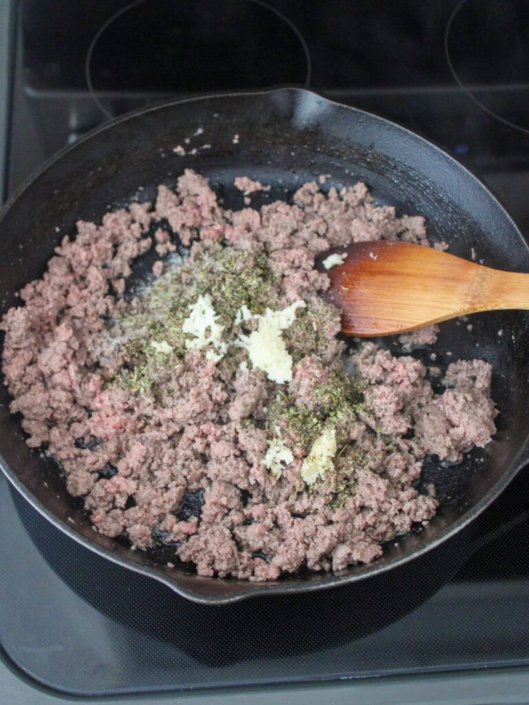 ground beef, salt, pepper, Italian seasoning, garlic in a cast iron skillet cooking with a wooden spoon