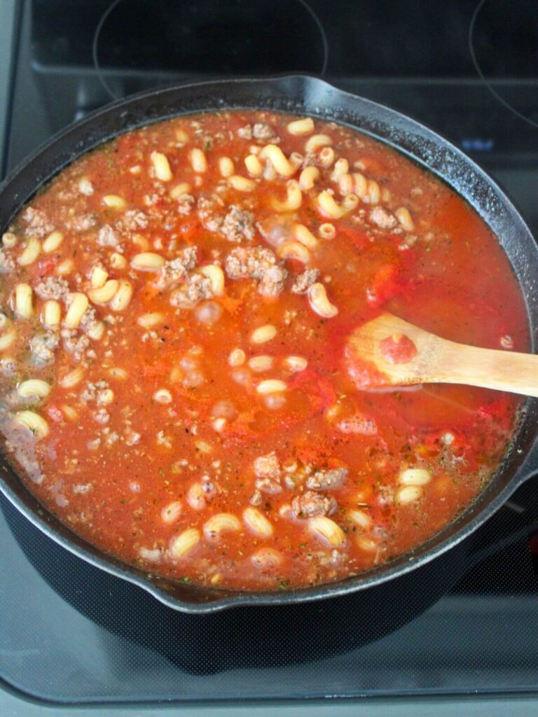 adding the bone broth, marinara and pasta to the ground beef mixture in the cast iron skillet with a wooden spoon