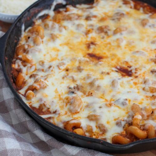 the pasta bake resting on a cutting board in the cast iron skillet with a plaid towel next to it