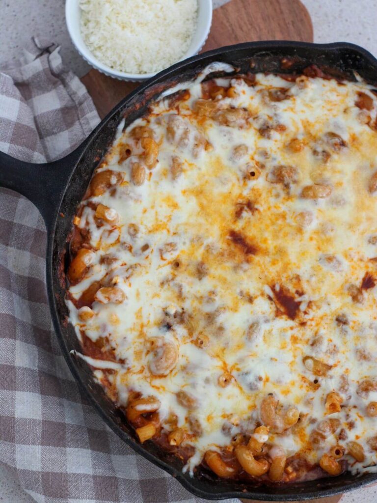 One-Pan Creamy Pasta bake ready to eat in a cast iron skillet on a cutting board with a plaid napkin and small white bowl of parmesan cheese