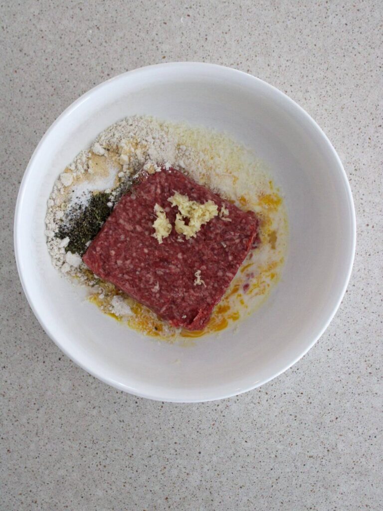 ground beef, egg, oats, salt, pepper, Italian seasoning, garlic, milk and parmesan in a white bowl ready to mix