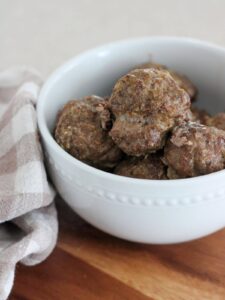 simple baked meatballs with oats in a white bowl on a brown cutting board with a plaid napkin next to it.