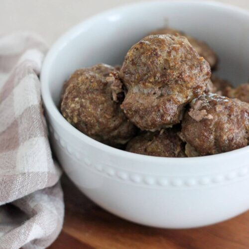 simple baked meatballs with oats in a white bowl on a brown cutting board with a plaid napkin next to it.