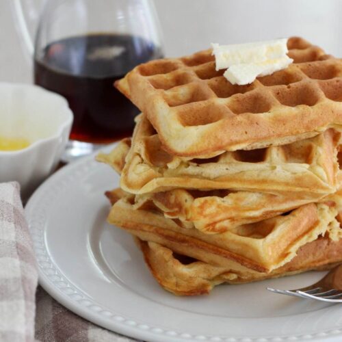 Sourdough discard waffles stacked on a white plate with butter on tip with maple syrup and melted butter in the back with a plaid napkin and silver spoon