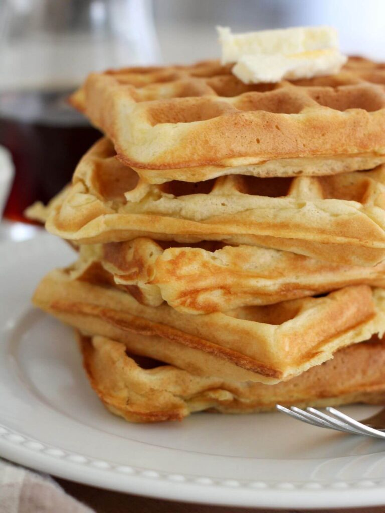 sourdough discard waffles stacked up high on a white plate with butter on top and a silver spoon