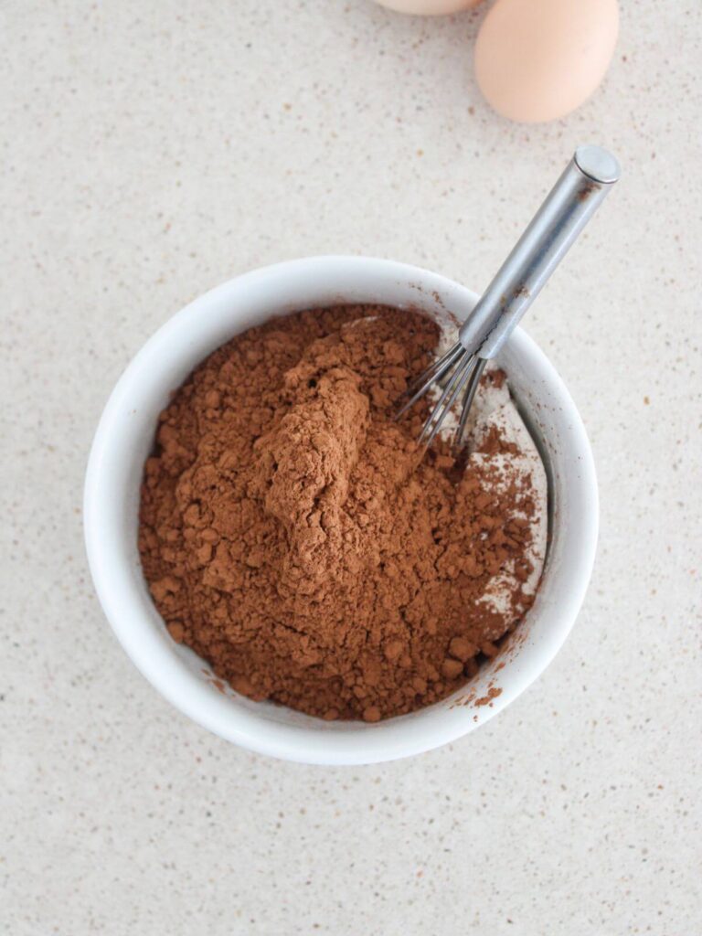 whisking the flour, cocoa powder, salt and baking soda in a white bowl with a metal whisk