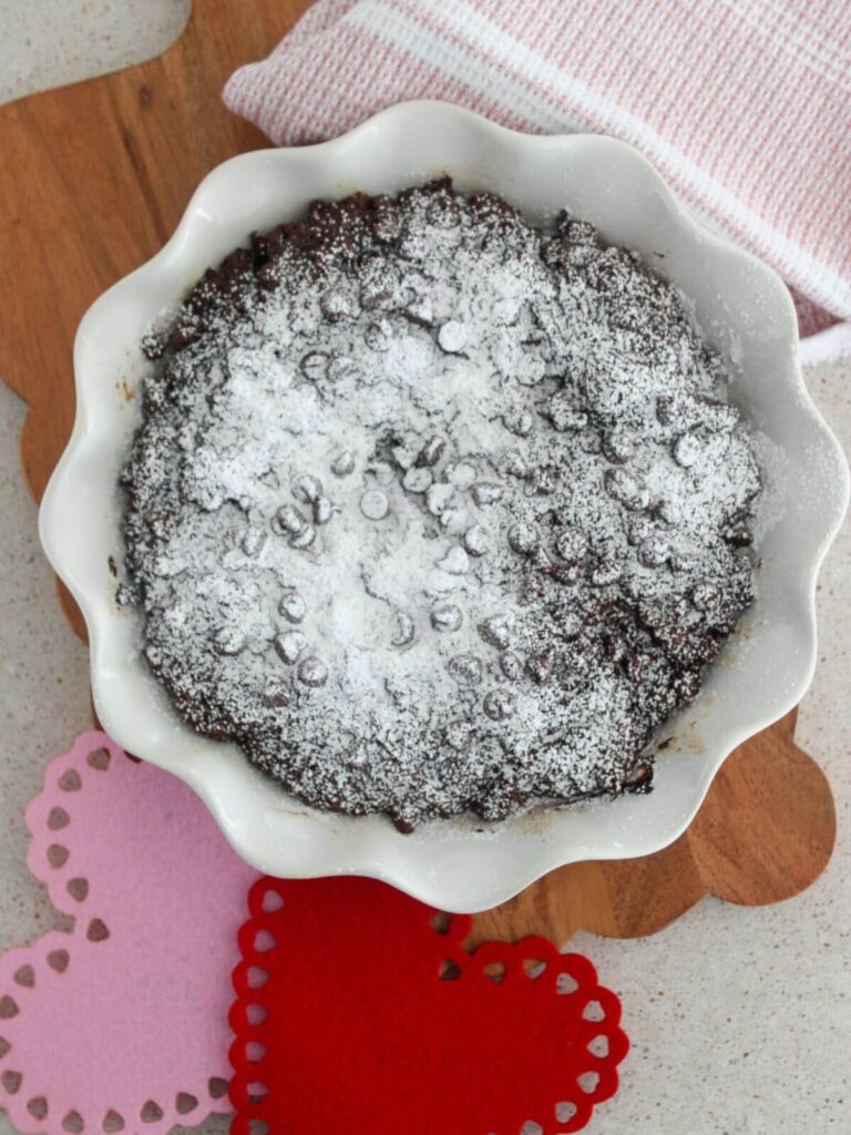 Valentine's Chocolate Snacking Cake baked in a white pie pan on a cutting board with a pink and red heart next to it and a pink and white towel next to it