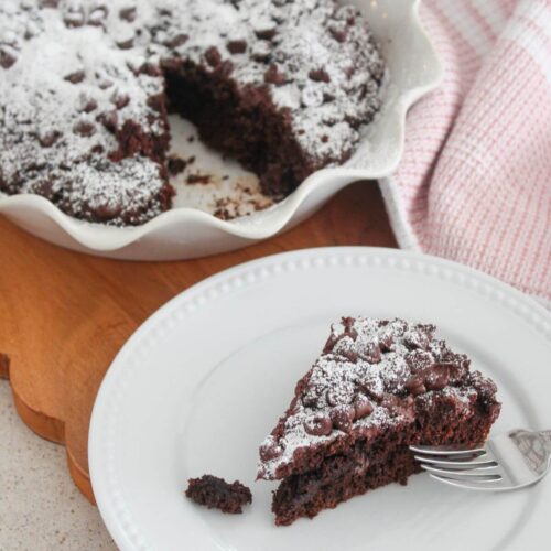 a slice of chocolate cake on a white plate with a spoon with the rest of the cake in a pie pan on a cutting board with a pink and white towel