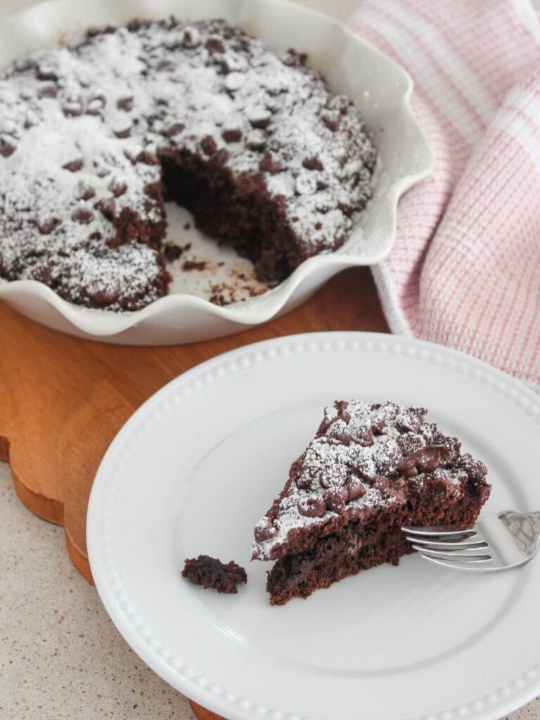 a slice of chocolate cake on a white plate with a spoon with the rest of the cake in a pie pan on a cutting board with a pink and white towel