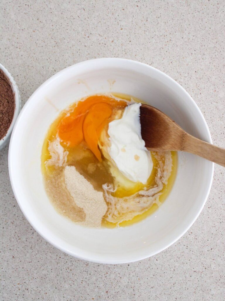 mixing the Greek yogurt, eggs, cane sugar, butter and vanilla in a white bowl with a wooden spoon and the dry mixture next to it