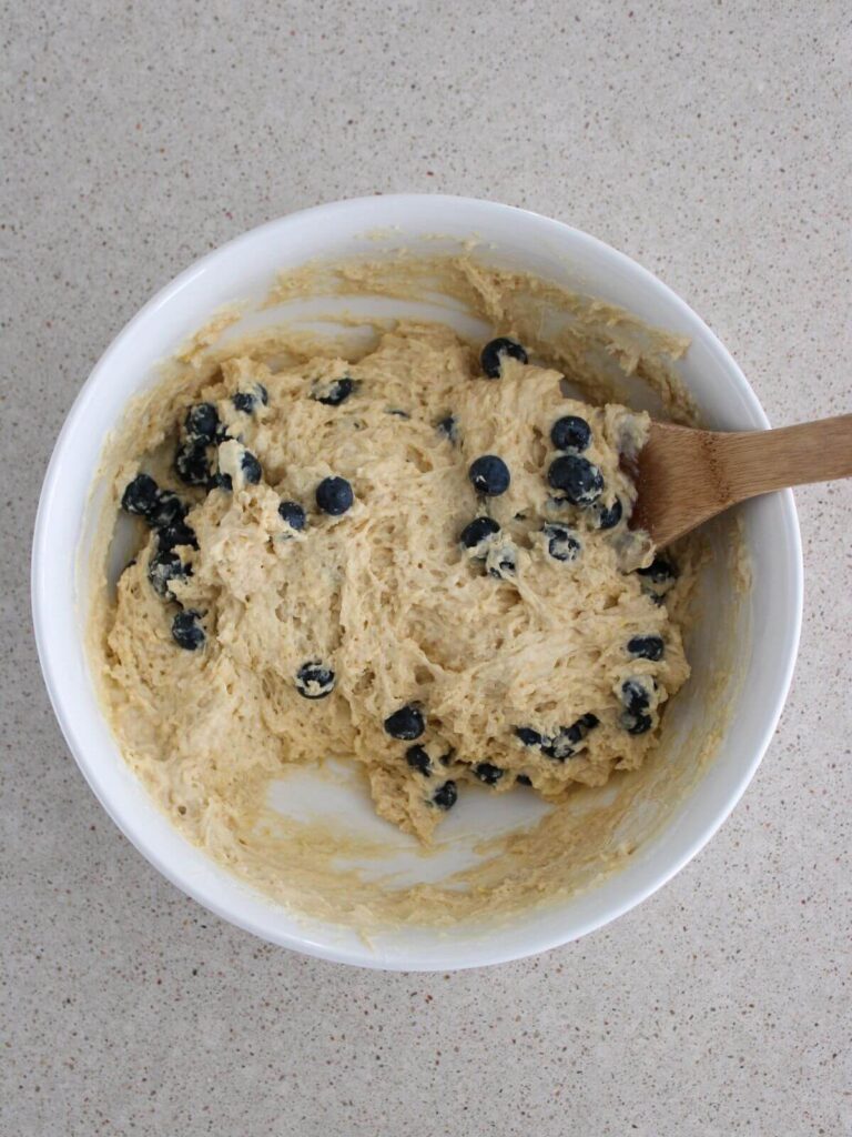 Mixing the blueberries into the dough in a white bowl with a wooden spoon