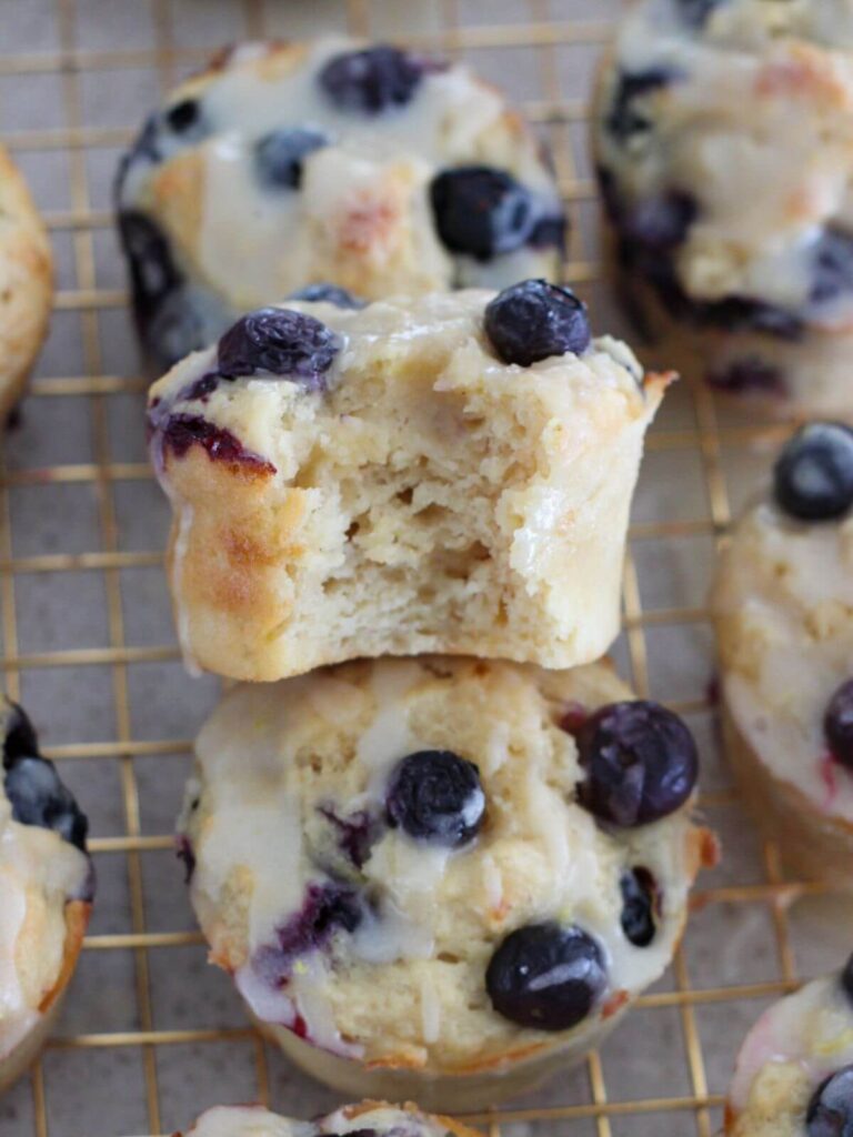 Blueberry Lemon Muffins stacked on top of others with a bite taken out and a golden cooling rack underneath