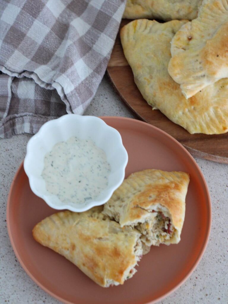 chicken bacon ranch pocket on a plate cut in half with a side cup of ranch with a plaid napkin and stack of more chicken bacon ranch pockets in the background