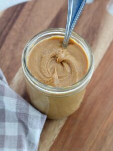 peanut butter in a glass mason jar with a silver spoon on a cutting board with a plaid napkin