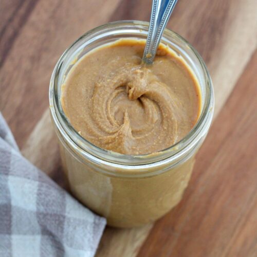 peanut butter in a glass mason jar with a silver spoon on a cutting board with a plaid napkin
