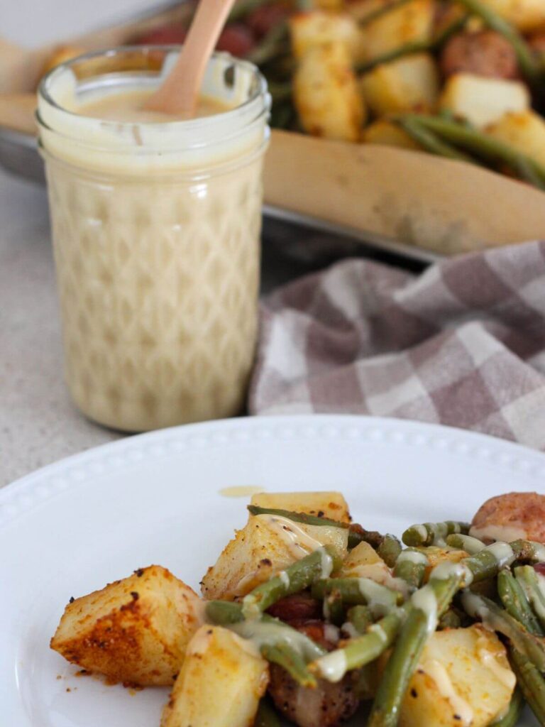 honey mustard in a mason jar with wooden spoon with a plate of sheet pan chicken sausage, potatoes and green beans in front of it with the sheet pan behind and a plaid napkin