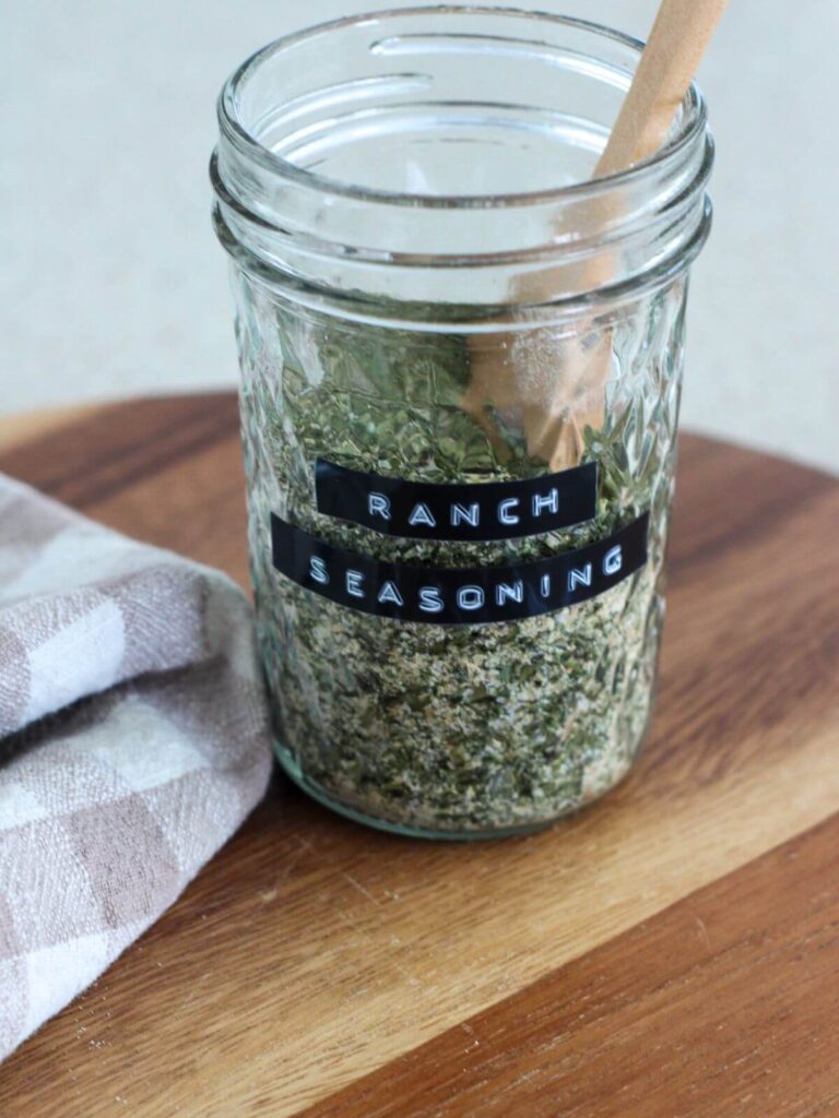 homemade ranch seasoning in a mason jar labeled ranch seasoning with a wooden spoon on a cutting board with a plaid napkin