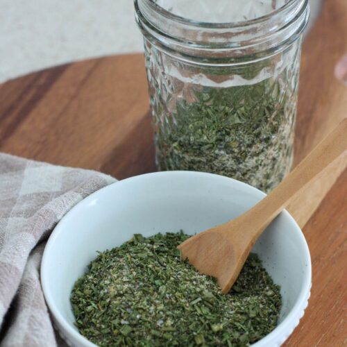 homemade ranch seasoning in a white bowl with a wooden spoon with the mason jar of seasoning behind with a plaid napkin next to it on a cutting board