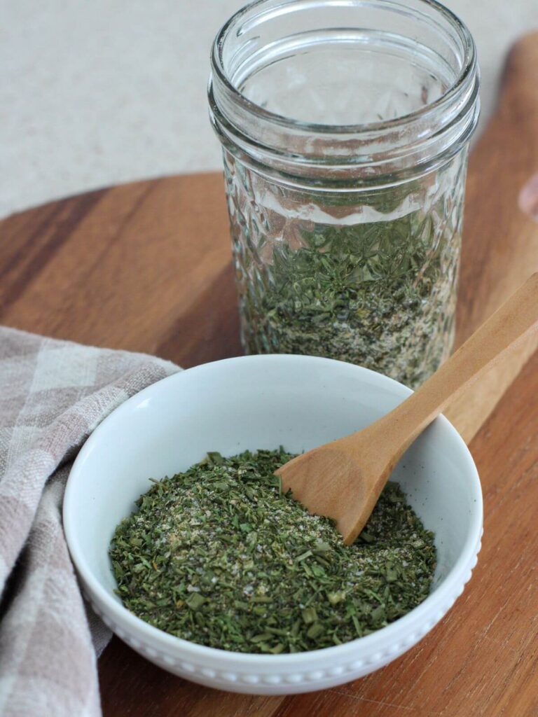 homemade ranch seasoning in a white bowl with a wooden spoon with the mason jar of seasoning behind with a plaid napkin next to it on a cutting board