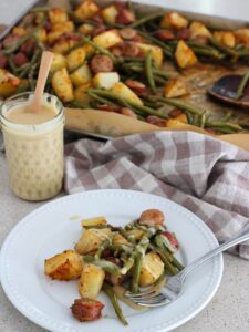 sheet pan chicken sausage on a white plate with a silver spoon, honey mustard in a mason jar and sheet pan in the back with a plaid napkin
