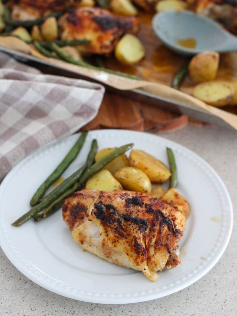 sheet pan lemon chicken and potatoes with green beans on a white plate with the rest of the sheet pan in the background along with a plaid napkin