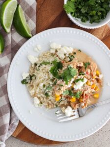 Slow Cooker Corn Chicken Plated on a white plate with a silver spoon sitting on a wooden cutting board with limes, cilantro in a white bowl and a plaid napkin around it