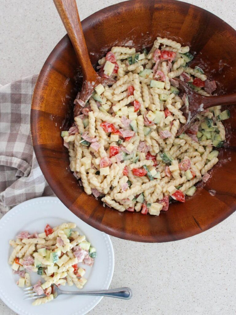 Pasta salad on a white plate with a silver spoon and the rest of the pasta salad in a big wooden bowl with a wooden spoon and a plaid napkin next to it